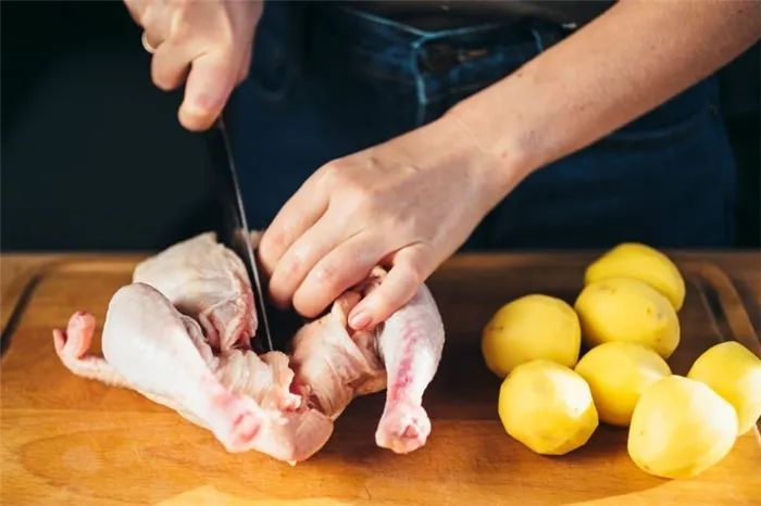 мясо цыпленка, которое разделывают на деревянной разделочной доске ножом, рядом лежит очищенная картошка