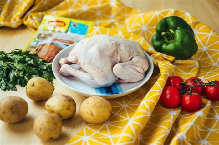 свежая тушка цыпленка в тарелке на столе, рядом лежит болгарский перец, помидоры, картошка, свежая зелень и пачка с приправой для курицы